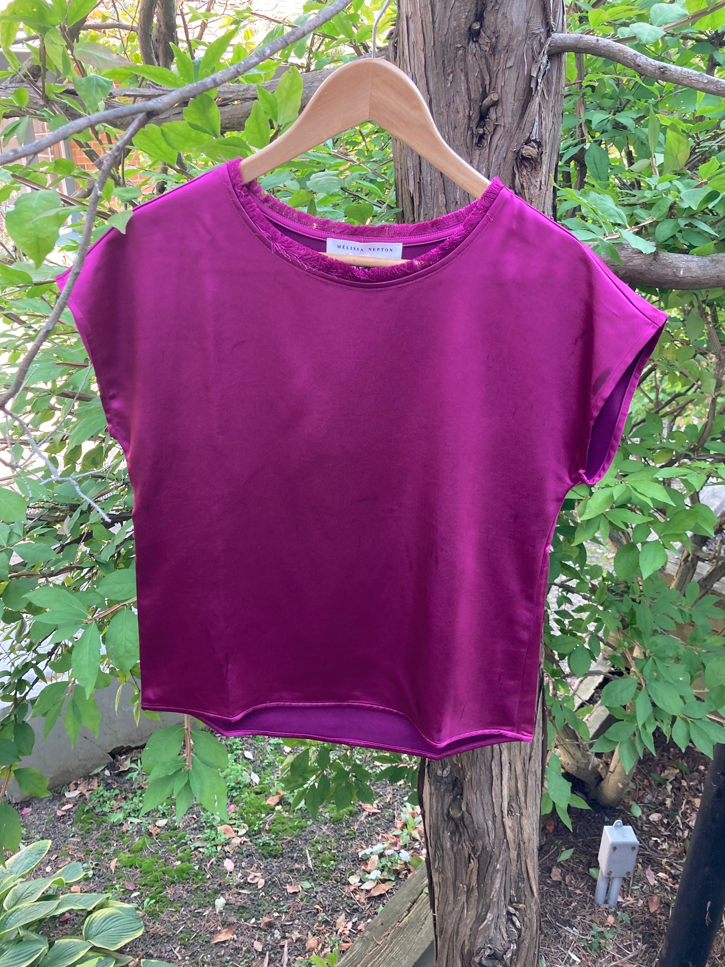 The willow top by Melissa Nepton hanging on a hanger in the woods, displaying the rough cut hem at the bottom and satin finish.
