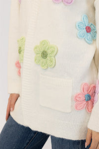 Close-up detail of the Elska cardigan’s textured flower embroidery in green, pink, and blue on soft white knit, showing pocket and ribbed hem.