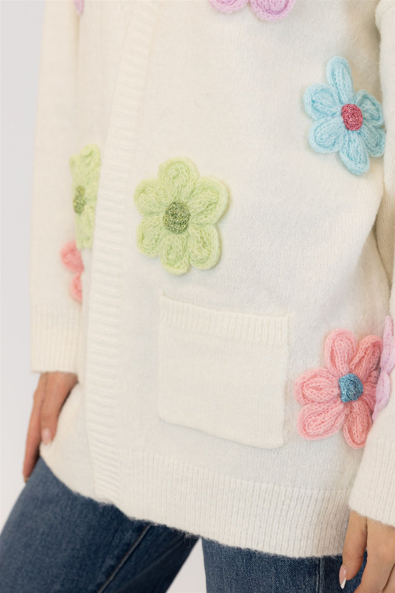 Close-up detail of the Elska cardigan’s textured flower embroidery in green, pink, and blue on soft white knit, showing pocket and ribbed hem.