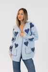 Main image: Woman wearing pale blue Lyla & Luxe ELSKAHEART cardigan with navy hearts, standing against white background