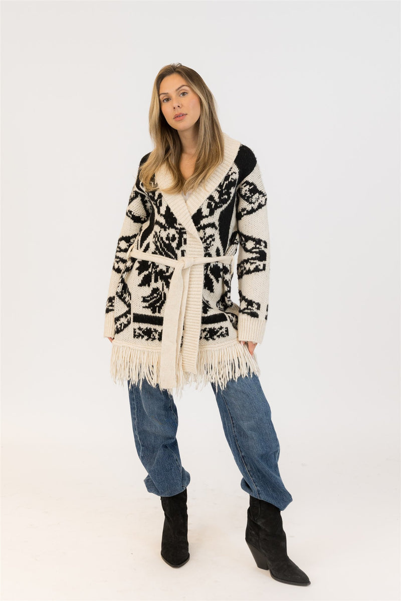 Woman wearing the Lyla + Luxe Zane cardigan in beige and black, standing front-facing with hands tucked in the pockets, styled with slouchy jeans and black heeled boots — photographed against a white studio backdrop. 