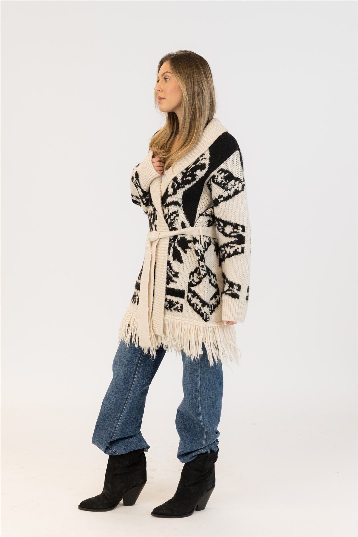 Side profile of model in the Zane cardigan showing the shawl collar, belted waist, and bold knit pattern — styled with denim and suede boots.

