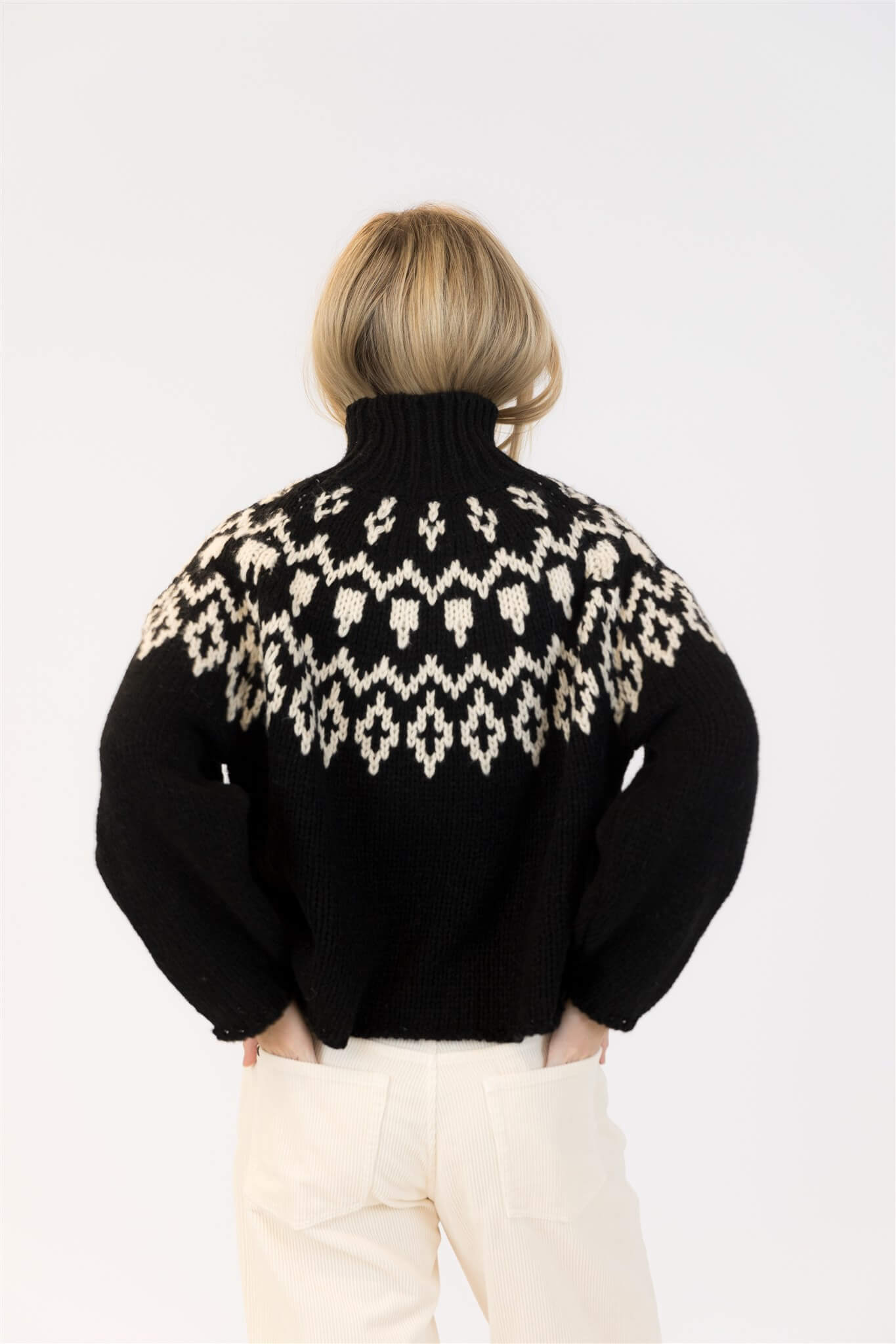 Back view of Lyla & Luxe Aggie Fair Isle turtleneck sweater in black and white, styled with cream pants.

