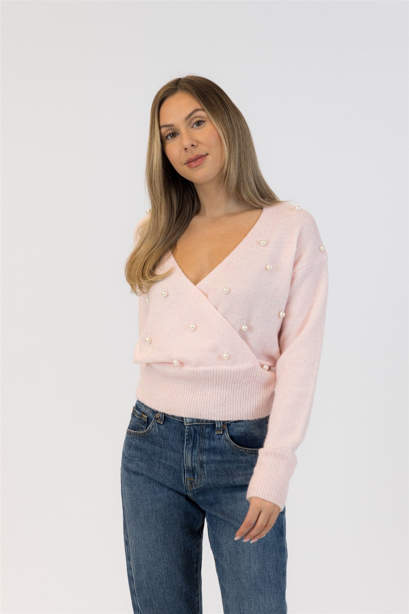 Woman wearing a light pink sweater with pearl embellishments and blue jeans on a white background