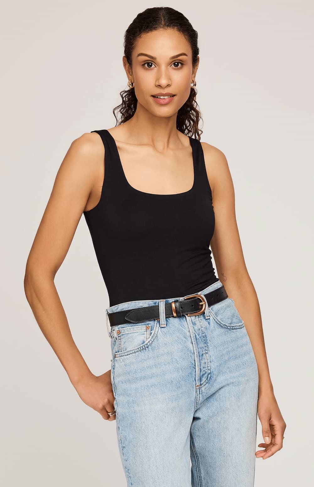 A woman modeling a black scoop-neck tank top paired with light-wash blue jeans and a black belt, photographed against a plain light gray studio background.