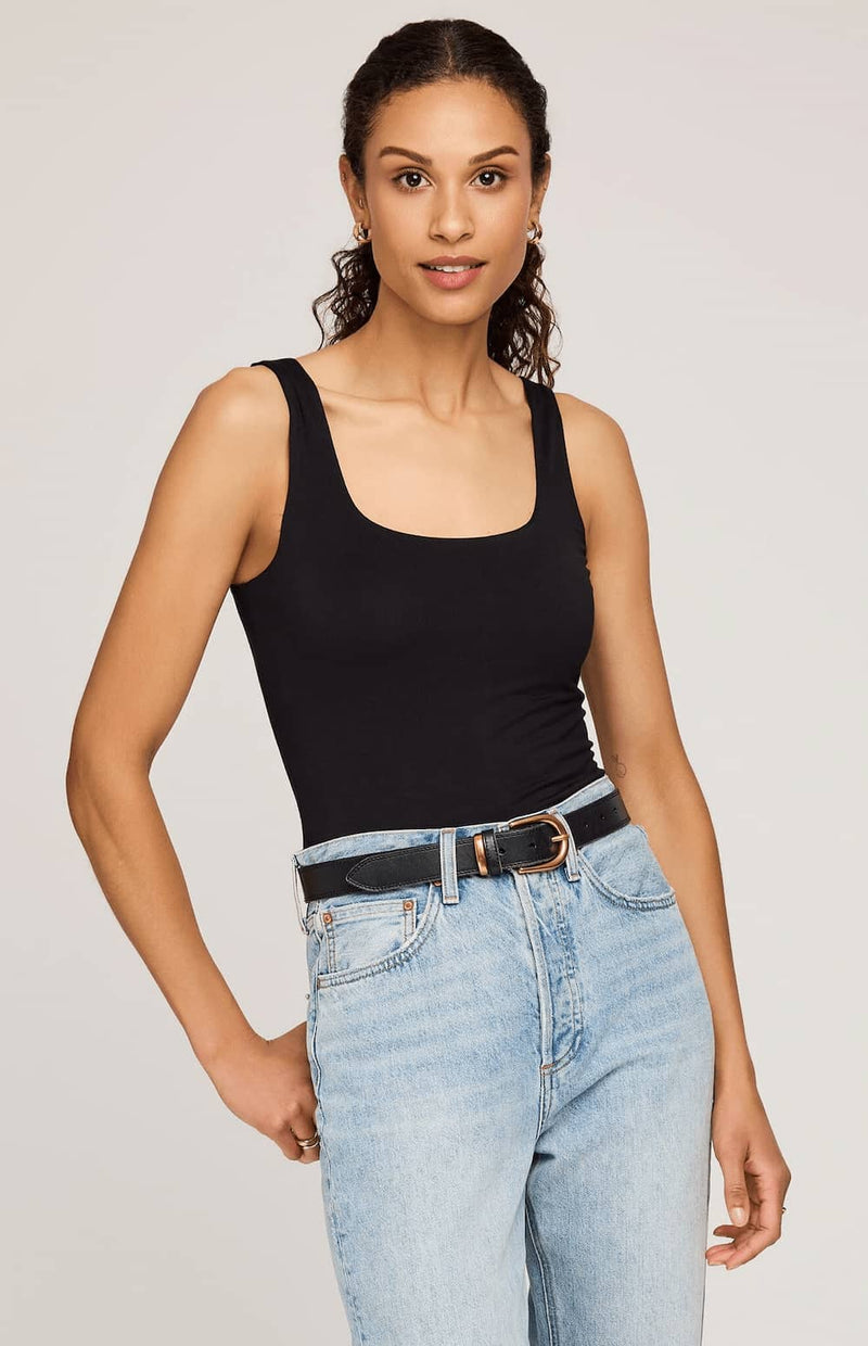 A woman modeling a black scoop-neck tank top paired with light-wash blue jeans and a black belt, photographed against a plain light gray studio background.