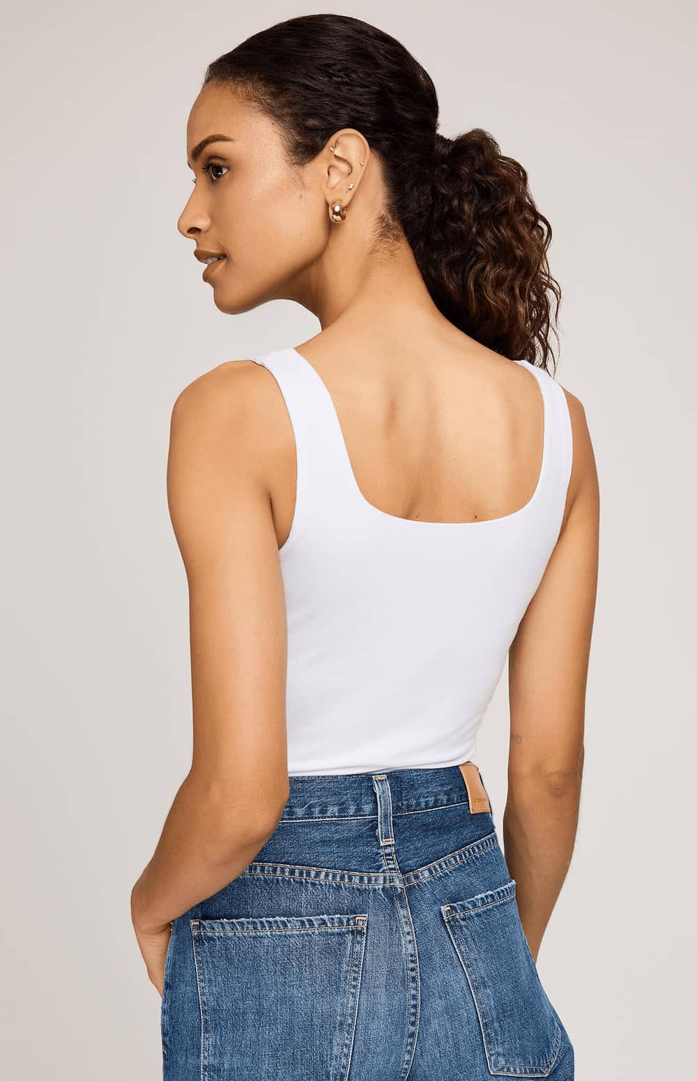 A woman shown from the back wearing a white scoop-neck tank top and blue jeans against a light gray studio background.