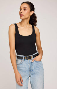 A woman modeling a black scoop-neck tank top paired with light-wash blue jeans and a black belt, photographed against a plain light gray studio background.