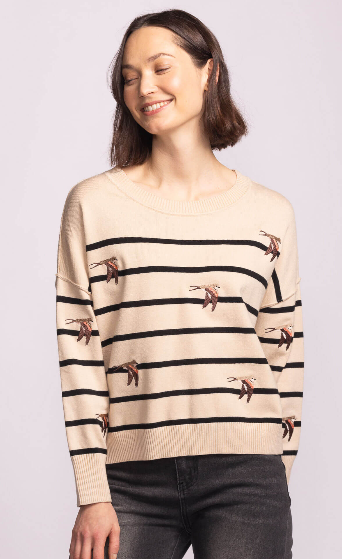 Model smiling in the Cruz Sweater, beige striped knit with whimsical swallow embroidery, casual chic layering piece.