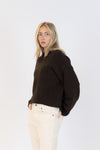 Side angle of model wearing a slouchy brown EVA Luxe knit sweater and cream pants, looking directly at camera.