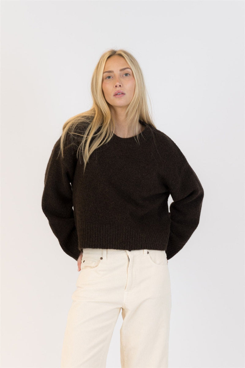 Blonde model wearing the chocolate EVA Luxe sweater from Lyla + Luxe with hands behind her back, styled casually with high-waisted ivory pants.