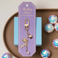 Gold Bella Bag Charm styled on lavender card, displayed with disco ball accents – Foxy Originals at Lemonberry