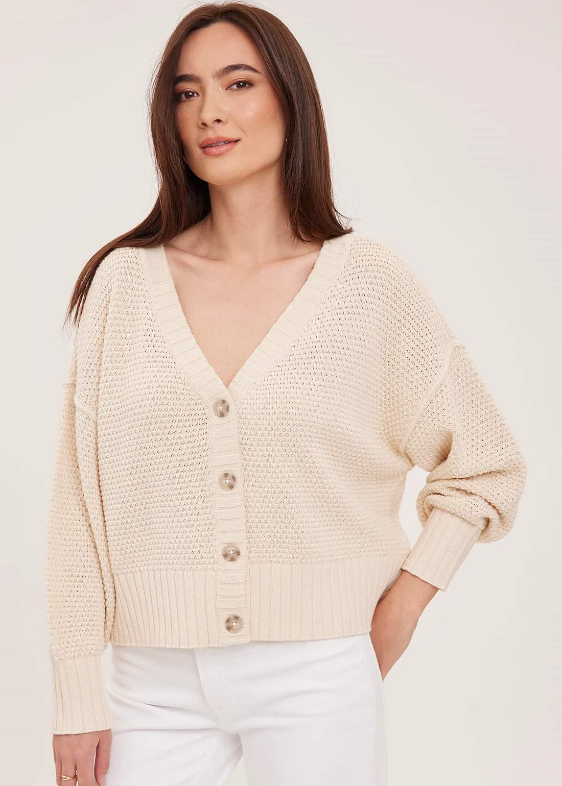 A woman wearing a cream-colored, textured knit cardigan with a V-neck and button-front closure, paired with white pants, against a plain light background.