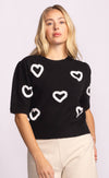 Close-up of black knit sweater with bold white heart design, styled with beige trousers.