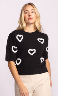 Close-up of black knit sweater with bold white heart design, styled with beige trousers.