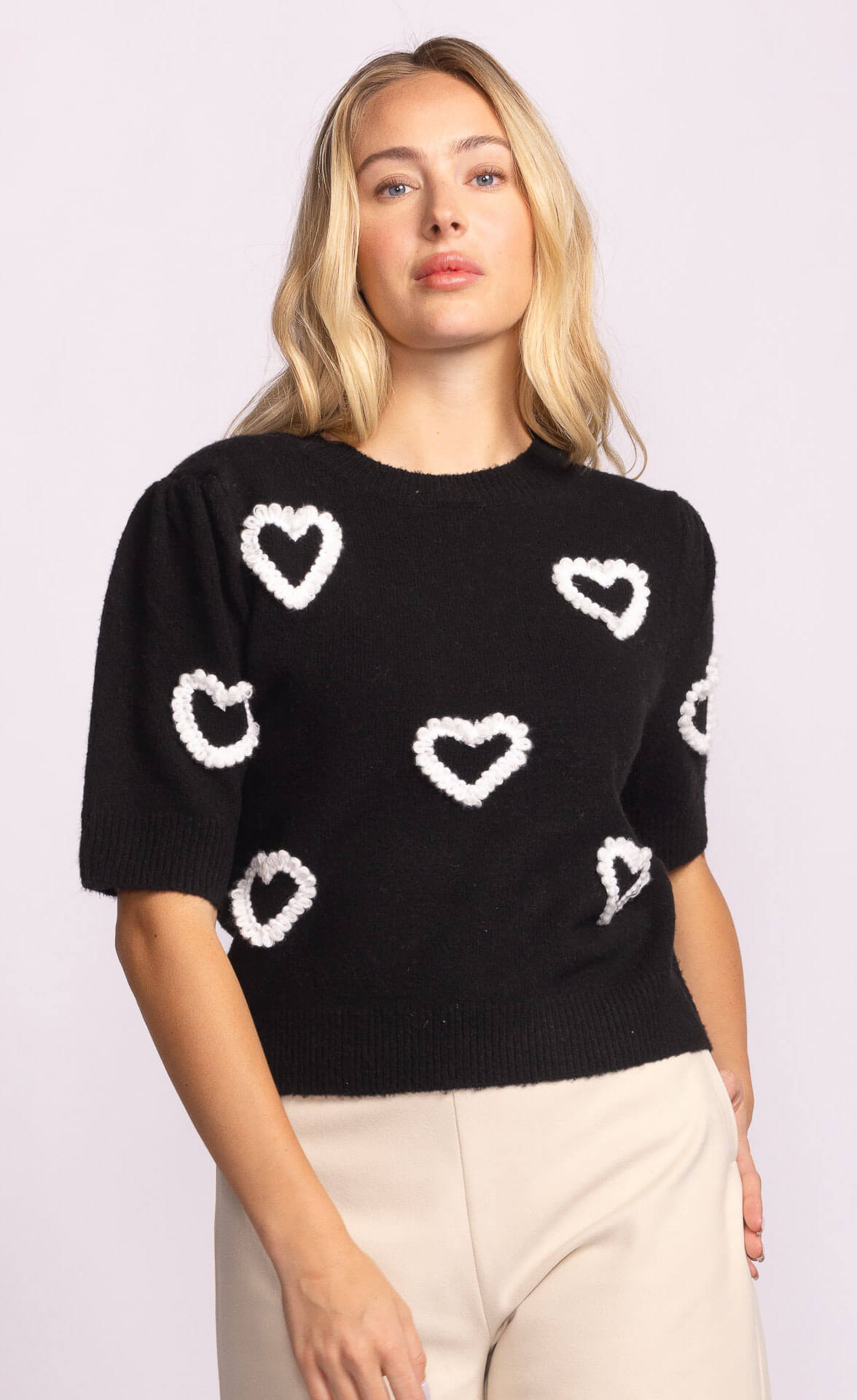 Close-up of black knit sweater with bold white heart design, styled with beige trousers.