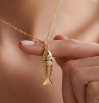 Gold fish-shaped pendant necklace held by a hand against a neutral background