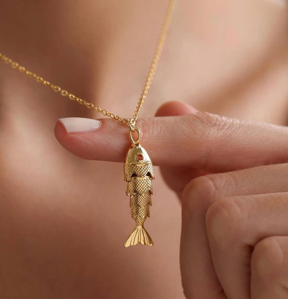 Gold fish-shaped pendant necklace held by a hand against a neutral background