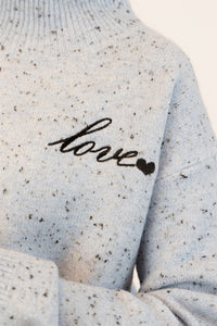 Closeup details of Lyla & Luxe's speckled gray sweater with 'love' embroidery on a neutral background