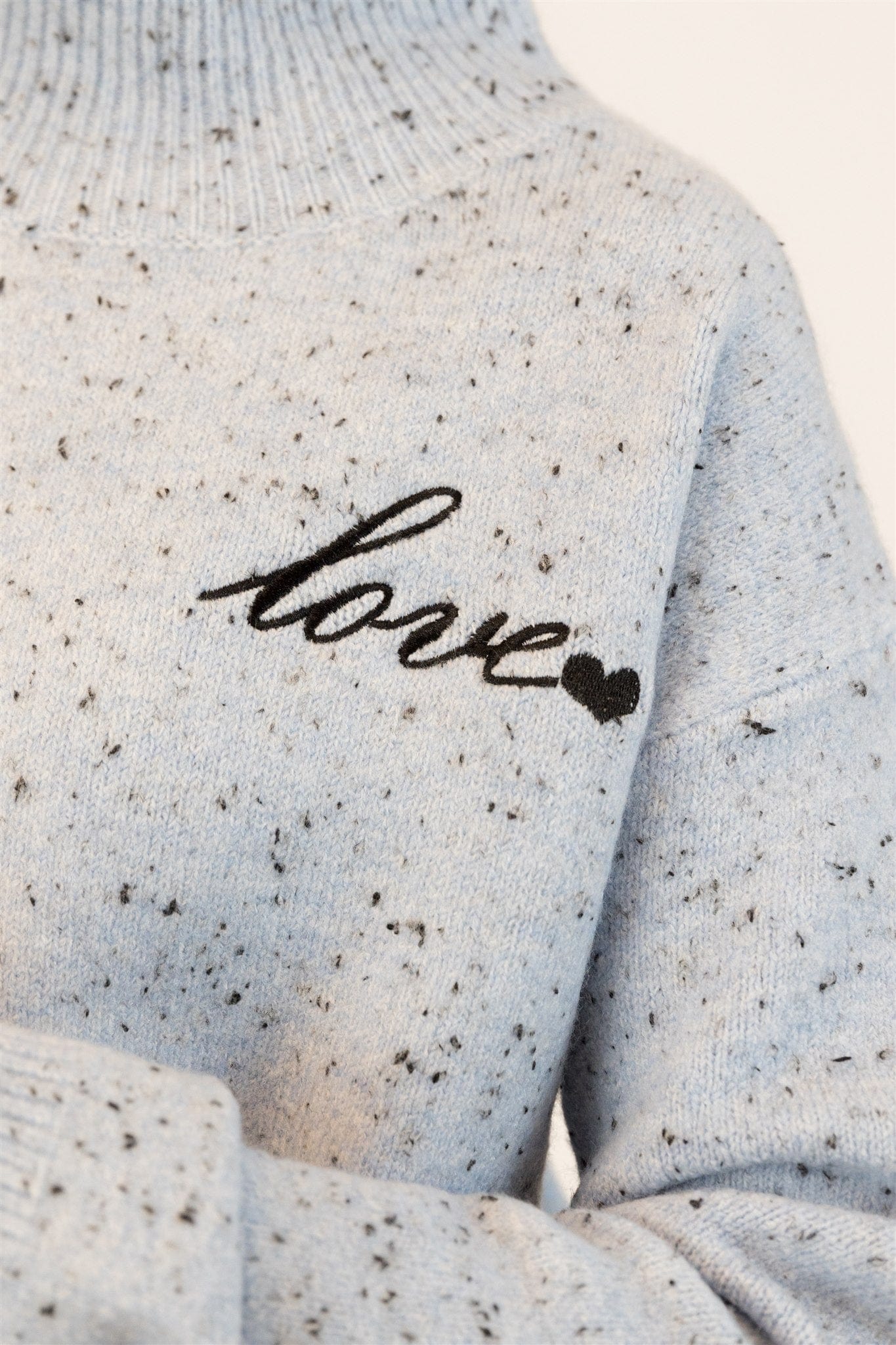 Closeup details of Lyla & Luxe's speckled gray sweater with 'love' embroidery on a neutral background