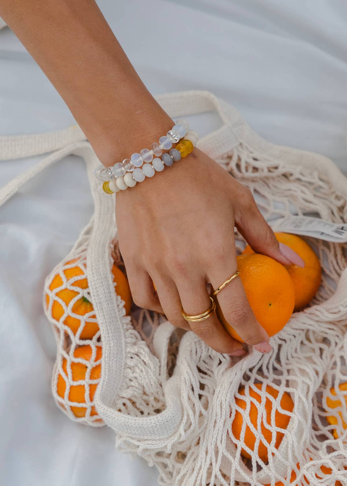 Stacked Namaste Jewellery Dania Stone Bracelets with clear and white beads worn with gold rings beside a market bag of oranges