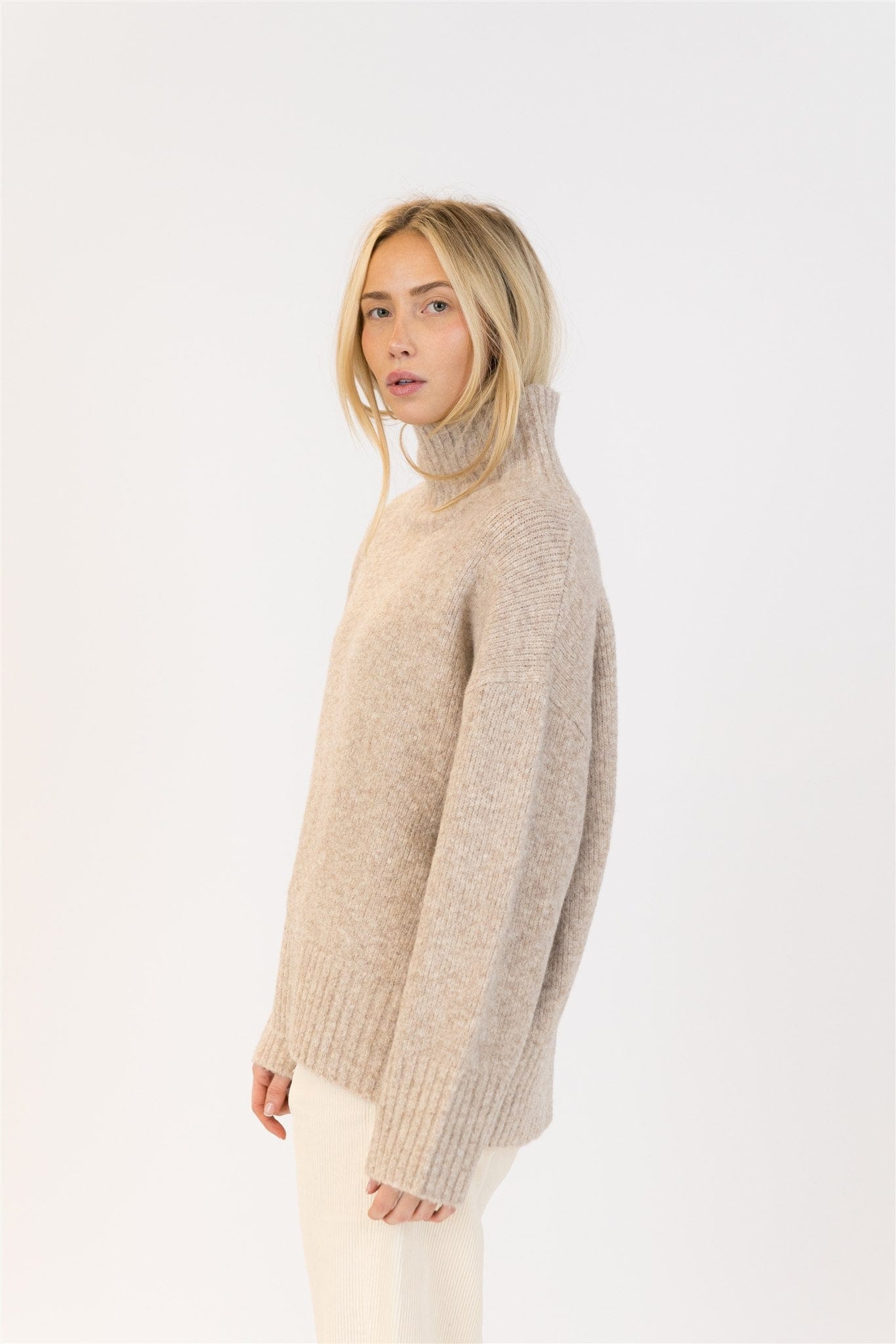 Side view of mockneck sweater in oat, styled with soft waves and minimalist cords