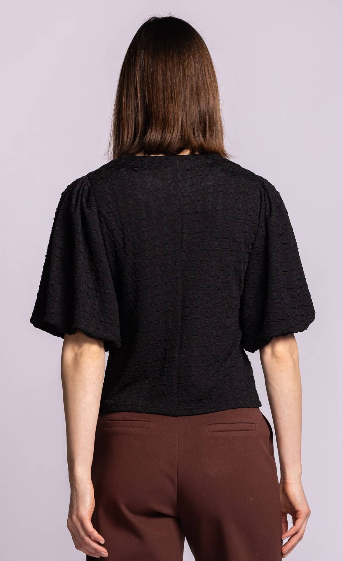 A woman viewed from the back wearing a black textured short-sleeve top and brown pants against a light lavender studio background.