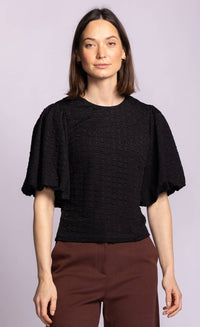 A woman modeling a black textured short-sleeve top with dramatic flutter sleeves and brown trousers, set against a solid light purple background.