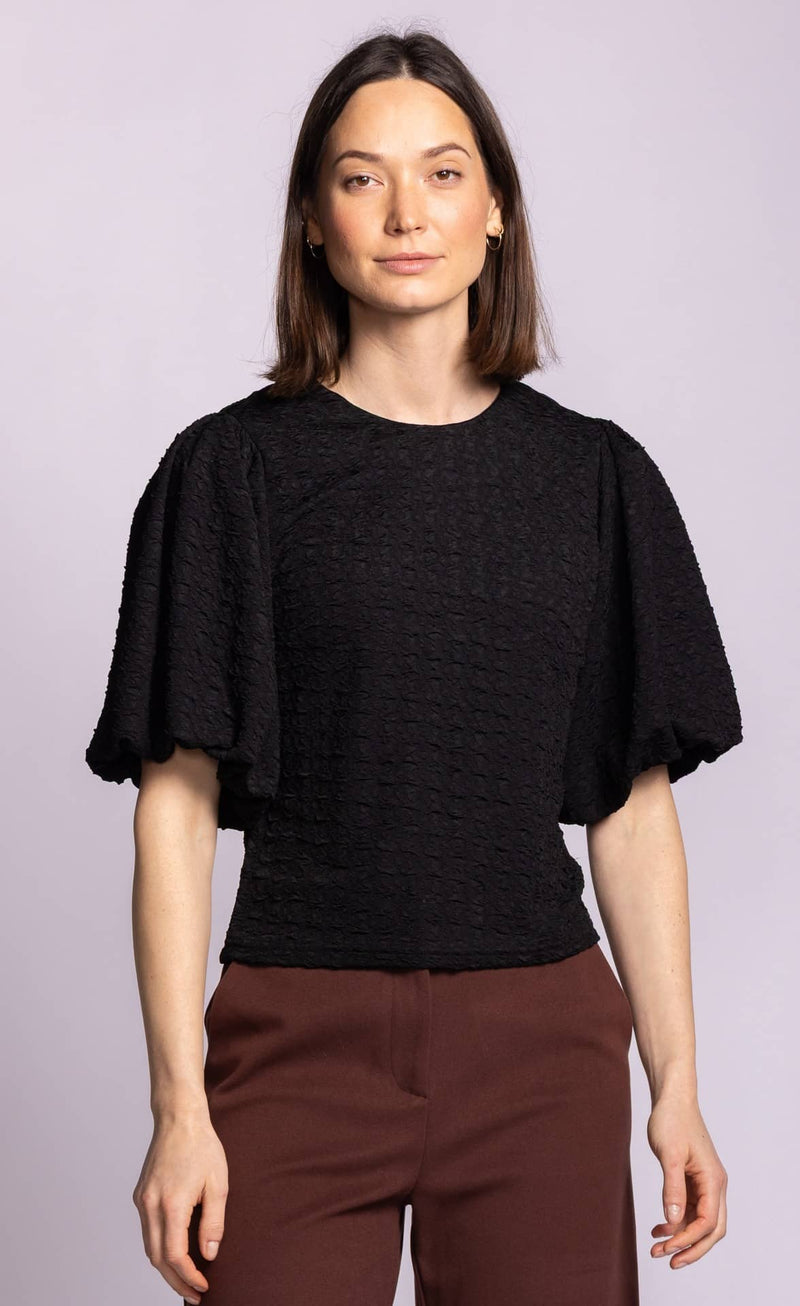 A woman modeling a black textured short-sleeve top with dramatic flutter sleeves and brown trousers, set against a solid light purple background.