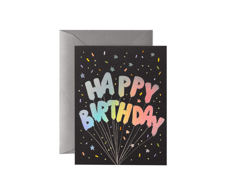 Rifle Paper Co. birthday card with rainbow foil balloon letters on black background, styled with silver envelope.