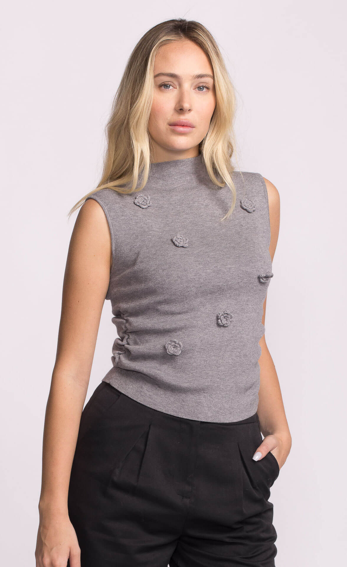 Model wearing grey sleeveless knit Rosslie Sweater with mock neck and floral appliqué detail, styled with black pants.