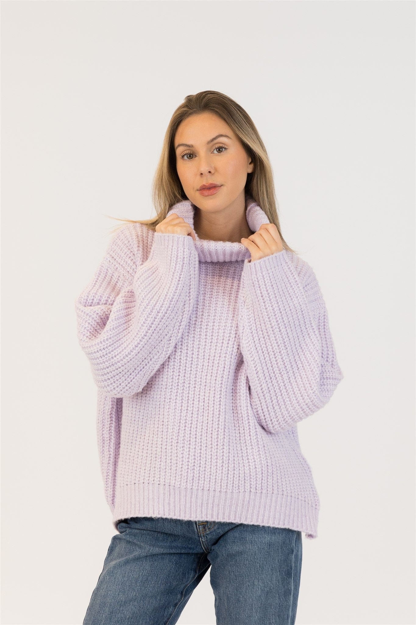 Model wearing lilac Lyla + Luxe SAHAR sweater facing front, holding ribbed turtleneck with both hands, styled with blue jeans, white backdrop