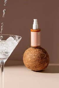 St. Barts dry shimmering oil in bronze balanced on a coconut, photographed with a cocktail glass, available at Lemonberry Aurora.