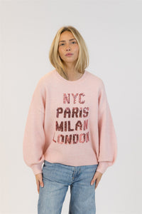 Front view of model wearing Lyla + Luxe Yassie sequin sweater in soft pink with city names in rose-gold sequins, photographed on white background