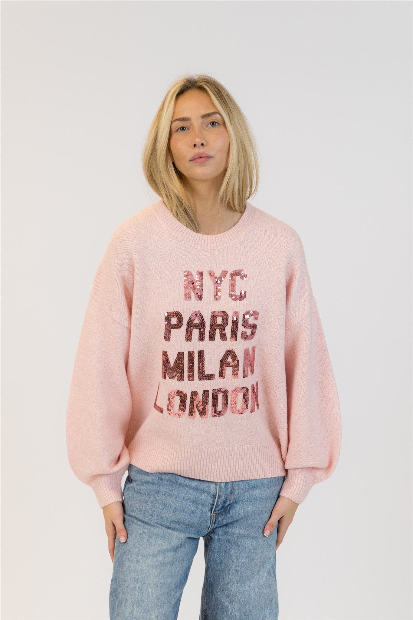 Front view of model wearing Lyla + Luxe Yassie sequin sweater in soft pink with city names in rose-gold sequins, photographed on white background